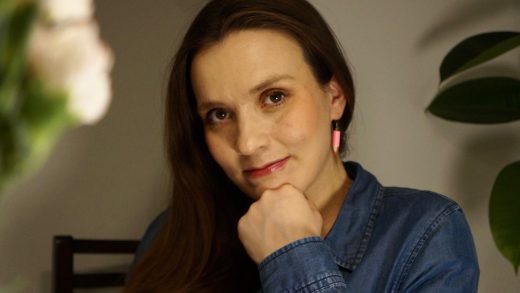 Nainen, jolla on pitkät ruskeat hiukset, farkkupaita ja vaaleanpunaiset korvakorut, nojaa leukansa käteensä ja hymyilee kevyesti istuessaan sisätiloissa, ja etualalla ja taustalla on hämäriä kasveja.