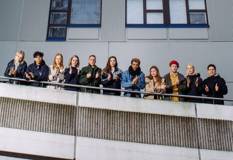 Kaksitoista ihmistä seisoo vierekkäin parvekkeella, kasvot eteenpäin ja taputtavat, taustalla rakennus, jossa on ikkunoita ja vaakasuora sivuraide.