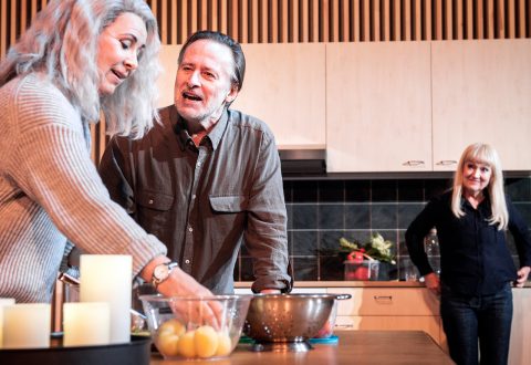 Kolme aikuista on modernissa keittiössä. Harmaahiuksinen nainen seisoo tiskin ääressä valmistelemassa ruokaa, mies kumartuu eteenpäin ja puhuu vilkkaasti, ja toinen nainen seisoo taustalla tarkkailemassa heitä ja hymyilee.