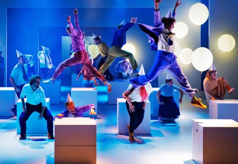 Värikkäisiin asuihin ja juhlahattuihin pukeutuneet esiintyjät hyppivät ja poseeraavat tarmokkaasti lavalla suurten valkoisten laatikoiden ja hehkuvien pyöreiden valojen ympäröimänä, mikä luo vilkkaan ja dynaamisen kohtauksen sinisellä ja lämpimällä valaistuksella.