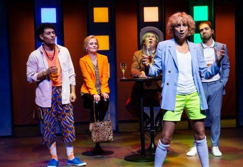 Viisi värikkäisiin, eklektisiin asuihin pukeutunutta ihmistä seisoo ja istuu lavalla, jossa on elävä valaistus. Yksi edessä oleva henkilö näyttää puhuvan intohimoisesti juoma kädessään; muut katselevat vaihtelevin ilmein.