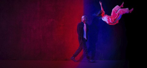 Tummiin vaatteisiin pukeutunut mies seisoo ja katsoo valkoiseen mekkoon pukeutunutta naista, joka näyttää leijuvan tai putoavan ilmassa. Kohtaus on valaistu dramaattisella punaisella ja sinisellä valaistuksella, joka jakaa taustan kahteen väriin.