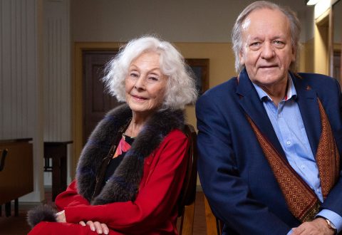 Vanhempi nainen, jolla on valkoiset hiukset ja punainen asu, istuu siniseen pukutakkiin ja siniseen paitaan pukeutuneen vanhemman miehen vieressä. He ovat sisätiloissa ja hymyilevät lempeästi kameralle.