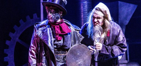 Kaksi näyttelijää näyttämöllä dramaattisissa puvuissa; toisella on kaasunaamari, silinterihattu ja nahkatakki sekä punainen huivi, kun taas toisella on pitkä valkoinen parta, yömyssy ja kaapu, ja hänellä on sauva kädessään. Sininen valaistus viittaa teatterilliseen steampunk-kohtaukseen.