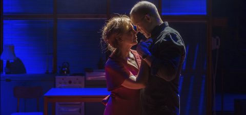 Mies ja nainen seisovat lähekkäin ja jakavat intiimin hetken dramaattisessa sinisessä ja violetissa näyttämövalaistuksessa, ja taustalla näkyvät keittiökalusteet ja kaihtimet.