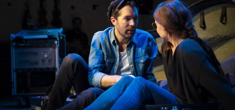 Mies ja nainen istuvat lattialla vastakkain ja keskustelevat näyttämövalaistuksessa. Heidän takanaan näkyy musiikkilaitteita, jotka viittaavat kulissien takaiseen tai esitysympäristöön.