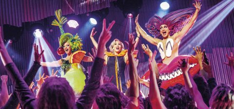 Lavalla on kolme värikkäisiin, taidokkaisiin pukuihin pukeutunutta drag-esiintyjää, jotka laulavat ja nostavat käsiään, kun innostunut yleisö kohotetuin käsin hurraa kirkkaiden valojen alla.