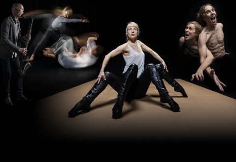 Dramaattinen kohtaus, jossa vasemmalla on saksofonisti, epäselviä dynaamisia tanssijoita keskellä ilmaa ja kolme esiintyjää, jotka ovat terävästi fokusoituja: istuva nainen mustissa saappaissa ja kaksi ilmeikästä paidatonta miestä hänen oikealla puolellaan, kaikki tummaa taustaa vasten.