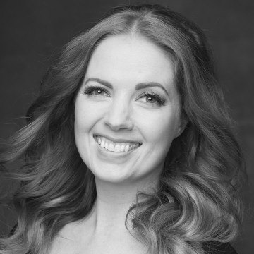 Mustavalkoinen muotokuva naisesta, jolla on pitkät, aaltoilevat hiukset ja joka hymyilee leveästi ja katsoo hieman sivulle. Tausta on tavallinen ja tumma, mikä korostaa naisen ilmeikkäitä kasvoja ja kirkasta hymyä.