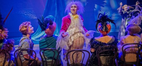 Kirkkaan vaaleanpunaiseen takkiin ja valkoiseen röyhelöpaitaan pukeutunut henkilö esiintyy näyttämöllä, jota ympäröivät värikkäisiin pukuihin ja höyhenpeitteisiin hattuihin pukeutuneet istuvat näyttelijät dramaattisen valaistuksen ja sinisen taustan alla.