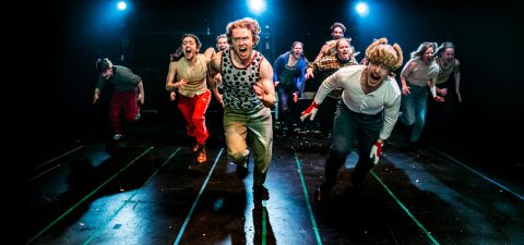 Ryhmä värikkäisiin pukuihin pukeutuneita esiintyjiä juoksee energisesti kohti kameraa pimeällä, kirkkaasti valaistulla näyttämöllä, ja dramaattiset ilmeet ja teatterivalaistus luovat kiireen ja jännityksen tunteen.