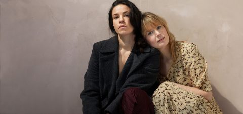 Kaksi naista istuu tiiviisti yhdessä beigeä seinää vasten; toisella on tumma takki ja viininpunaiset housut, kun taas toisella on vaalea kukkamekko ja hän nojaa kevyesti ensimmäisen naisen olkapäähän.