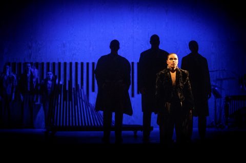 Henkilö seisoo dramaattisessa valaistuksessa hämärästi valaistun näyttämön edessä, ja taustalla on neljä varjohahmoa ja muusikoita, jotka kaikki kylpevät sinisessä valossa.