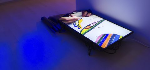 Lattialle sijoitettu suuri digitaalinen näyttö näyttää videon, jossa käsi piirtää värikkäitä abstrakteja muotoja. Osa kuvaruudusta on peitetty osittain rullattuun mustaan lakanaan, ja sininen valo loistaa lattian poikki.