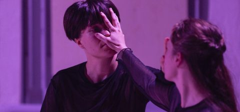 Kaksi tummiin vaatteisiin pukeutunutta ihmistä kohtaa toisensa violetissa valaistuksessa; toinen peittää toisen kasvot kädellään, mikä luo dramaattisen ja intensiivisen hetken.