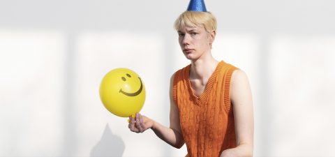 Siniseen juhlahattuun ja oranssiin hihaton puseroihin pukeutunut henkilö pitelee keltaista ilmapalloa, jossa on hymynaama. Henkilö näyttää vakavalta, mikä on ristiriidassa juhlallisten asusteiden kanssa.