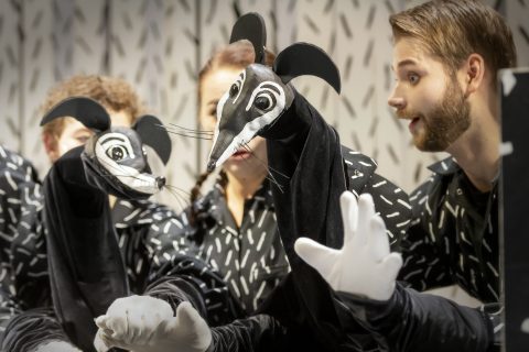 Kolme ihmistä käyttää suuria mustavalkoisia rotanukkeja, joilla on ilmeikkäät kasvot ja valkoiset hanskat, ja keskittyy keskittyneesti esitykseensä kuvioitua taustaa vasten.