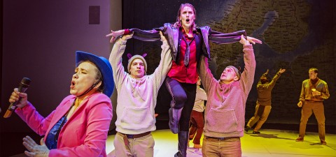 Vauhdikkaassa lavaesityksessä yksi henkilö laulaa mikrofoniin vaaleanpunaisessa bleiserissä ja sinisessä hatussa, kun muut nostavat dramaattista keskushahmoa kädet koholla. Taustalle heijastetaan kartta, ja muita näyttelijöitä seisoo lähellä.