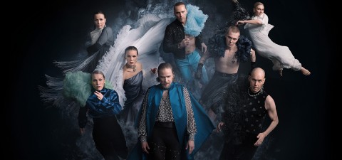 Yhdeksän tanssijan ryhmä, jolla on dramaattiset, virtaavat puvut, poseeraa dynaamisesti tummaa savutaustaa vasten luoden liikkeen ja energian tunteen.