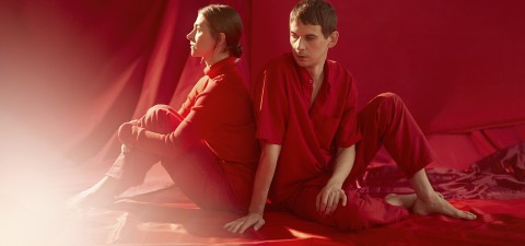 Punaiseen pukeutuneet nainen ja mies istuvat selkä toisensa vasten punaisella kangaslattialla punaisella taustalla, jota valaisee pehmeä, lämmin valo kuvan vasemmalta puolelta.