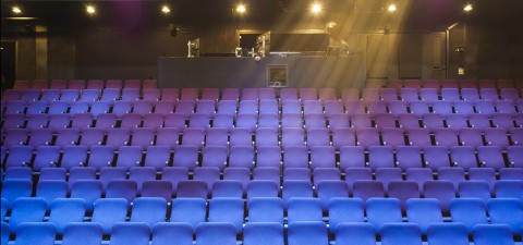 Tyhjiä violetteja teatteripenkkirivejä vastapäätä on takana oleva ohjauskoppi, joka on valaistu yläpuolella olevilla valoilla. Henkilö seisoo ohjauskopin takana, ja valonsäteet loistavat alas istuimille.