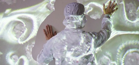 Valkoiseen laboratoriopukuun ja päähineeseen pukeutunut henkilö koskettaa seinää, kun abstrakteja, orgaanisia kuvioita heijastetaan sekä seinälle että hänen vartalolleen.