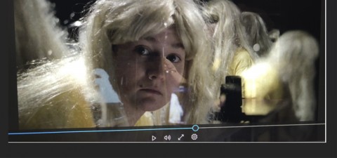 Henkilö, jolla on pitkä vaalea peruukki ja keltainen paita, katsoo kameraan, ja hänen heijastuksensa näkyy vieressä olevassa tummassa, lasimaisessa pinnassa. Videon toiston säätimet näkyvät kuvan alareunassa.