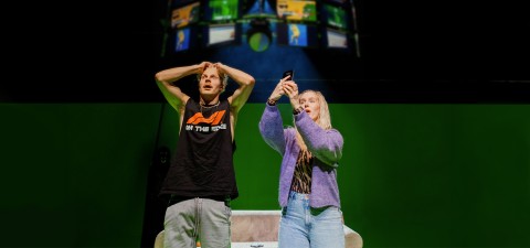 Kaksi ihmistä seisoo vihreän valkokankaan edessä; toinen pitää päätään järkyttyneenä, kun taas toinen nostaa puhelinta yllättyneen näköisenä. Heidän yläpuolellaan näkyy digitaalinen projektio, jossa on värikkäitä kuvia.