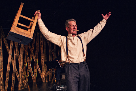 Mies seisoo näyttämöllä uhmakkaasti kädet ylhäällä pitäen toisessa kädessään tuolia. Taustalla on muusikko joka soittaa kitaraa.