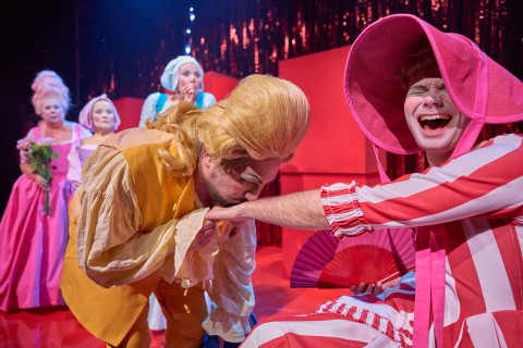 Teatterikohtaus, jossa näyttelijöillä on värikkäät, liioitellut aikakauden puvut. Keltaiseen pukeutunut mies kumartaa ja suutelee kättä toista, vaaleanpunaraitaiseen mekkoon ja suureen hattuun pukeutunutta miestä, joka nauraa, kun muut katselevat taustalla.