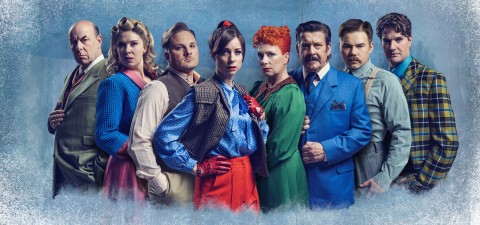 Kahdeksan värikkäisiin, vintage-henkisiin asuihin pukeutunutta ihmistä seisoo rivissä sinistä, kuvioitua taustaa vasten ja katsoo vakavana kameraan.