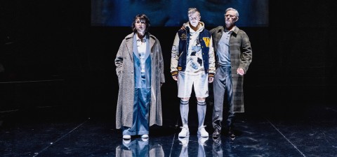 Kolme ihmistä seisoo valokeilassa tummaa taustaa vasten. Vasemmalla olevalla henkilöllä on pitkä takki ja vaaleansininen asu, keskellä olevalla henkilöllä on letterman-takki ja polvisukat, ja oikealla henkilöllä on silmälasit ja ruudullinen takki.