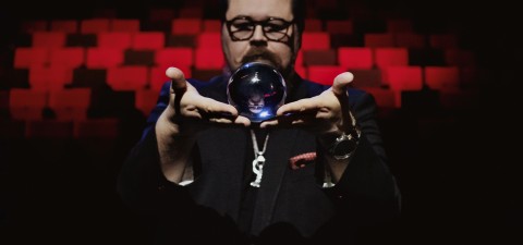 Silmälasipäinen, pukuun pukeutunut mies pitää kristallipalloa kätensä välissä, ja hänen takanaan on dramaattinen punainen ja musta tausta.