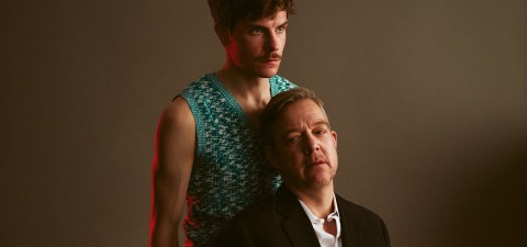 Nuori mies, jolla on kuvioitu hihaton paita, seisoo mustaan pukuun pukeutuneen vanhemman miehen takana. Molemmat katsovat vakavasti kameran ulkopuolelle neutraalia taustaa vasten, jossa on hienovarainen punainen valaistus.