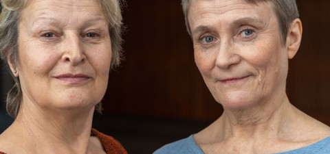 Kaksi vanhempaa naista, joilla on lyhyet harmaat hiukset ja rennot topit, seisovat lähekkäin ja katsovat kameraan lempein, neutraalein ilmein tummaa taustaa vasten.