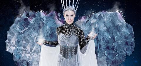 Jääkuningattareksi pukeutunut nainen seisoo kädet ojennettuina, yllään hopeinen kruunu ja koristeellinen jääpuku, jossa on kimaltelevia kristalleja ja huurteiset siivet tähtitaivaan taustalla.