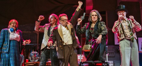 Lavalla seisoo viisi näyttelijää värikkäissä, omalaatuisissa puvuissa. Yksi nainen nostaa aseen, toinen hurraa, silmälasipäinen mies näyttää vakavalta, ja kaksi muuta reagoi dramaattisesti, mikä antaa kuvan vilkkaasta, teatraalisesta kohtauksesta.