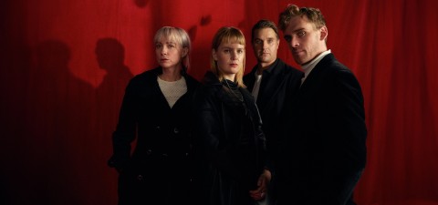 Neljä ihmistä seisoo punaisen verhon edessä tummiin, virallisiin vaatteisiin pukeutuneina. He katsovat kameraan vakavin ilmein, ja heidän varjonsa näkyvät heidän takanaan olevassa verhossa.