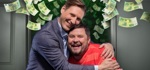 Kaksi hymyilevää miestä, toinen puvussa ja toinen punaraidallisessa paidassa, halaa toisiaan iloisesti, kun 100 euron seteleitä sataa heidän ympärilleen tummaa taustaa vasten.