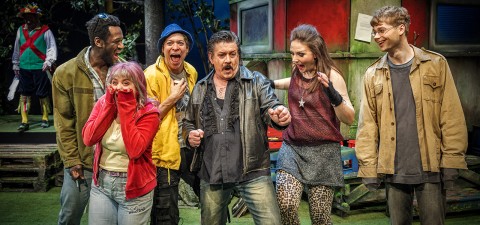 Kuusi ihmistä seisoo yhdessä ulkona ja ilmaisee yllätyksensä ja innostuksensa. Heillä on värikkäät, rennot vaatteet, ja he seisovat punaisen rakennuksen edessä, ja taustalla näkyy pukuun pukeutunut mies.