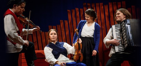 Lavalla on neljä muusikkoa, joilla on kansanomaiset vaatteet; yksi soittaa viulua, toinen pitelee viulua istuen, kolmas seisoo ja hymyilee, ja neljäs soittaa harmonikkaa. He esiintyvät puisen taustan edessä.