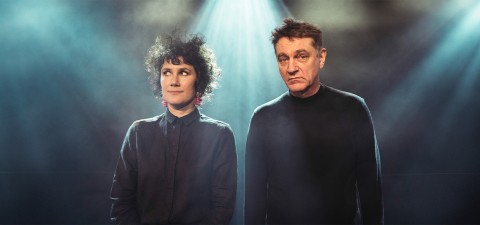 Kaksi ihmistä seisoo vierekkäin dramaattisen näyttämövalaistuksen alla. Vasemmalla olevalla henkilöllä on kiharat hiukset ja tumma paita sekä punaiset korvakorut, ja hän katsoo sivulle. Oikeanpuoleinen henkilö, jolla on lyhyet hiukset, katsoo eteenpäin, ja hänellä on yllään musta poolopaita.