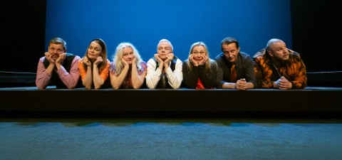 Seitsemän värikkäisiin vaatteisiin pukeutunutta ihmistä makaa lavalla vatsallaan, leuka kädessä ja hymyillen, sinistä taustaa vasten.