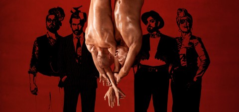 Kaksi paidatonta akrobaattia roikkuu ylösalaisin, koskettavat päitään ja tarttuvat käsillään toisiinsa punaisella taustalla, jossa on neljä mustavalkoista kuvitettua hahmoa, jotka ovat pukeutuneet erilaisiin pukuihin.