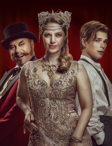 Punaisella taustalla poseeraa kolme ihmistä, joilla on taidokkaat, vintage-henkiset puvut: mies, jolla on silinterihattu ja punainen takki, nainen, jolla on jalokivikoristeinen mekko ja kruunu, ja nuori mies, jolla on valkoinen paita ja henkselit.