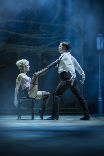 Korsettiin ja verkkosukkiin pukeutunut nainen istuu tuolilla ja ojentaa jalkansa koskettaakseen dramaattisesti seisovan smokkipukuisen miehen rintaa. Molemmat esiintyvät hämärästi valaistulla, savuisella lavalla.