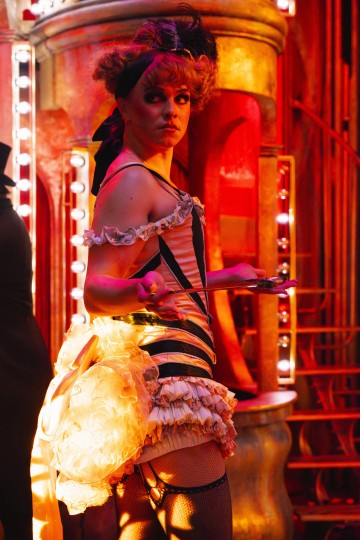 Nainen, jolla on röyhelöinen, raidallinen kabareeasu ja lavameikki, seisoo kirkkaasti valaistussa, punaisen ja kullanvärisessä teatterikuvassa, pitelee lautasia ja katsoo olkansa yli.