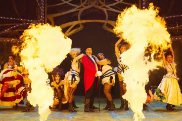 Punatakkinen sirkusjohtaja seisoo näyttämön keskellä, ja häntä ympäröivät burleskiasuihin pukeutuneet tanssijat, kun suuret liekit nousevat dramaattisesti molemmin puolin, ja taustalla on koristeltuja valoja ja lavasteita.