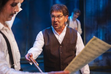 Liivi- ja silmälasipäinen mies elehtii intohimoisesti kapulalla ja puhuu samalla toiselle henkilölle, jolla on nuotit kädessään, ja taustalla sinisellä valaistulla näyttämöllä on kaksi muuta ihmistä.