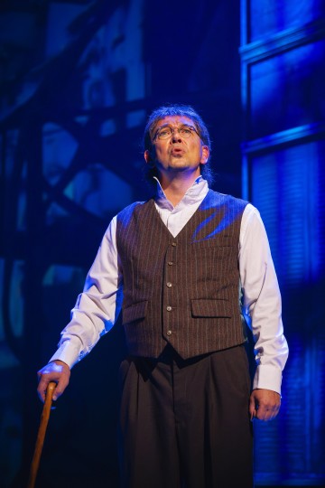 Mies, jolla on silmälasit, valkoinen paita ja ruskea neulenauhaliivi, seisoo näyttämöllä dramaattisen sinisessä valaistuksessa, pitää keppiä kädessään ja katsoo ylöspäin vakavalla ilmeellä.
